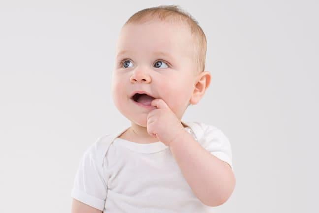 Smiling baby in a white outfit with a finger in their mouth, looking slightly to the side against a plain background